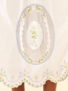 Close-up of Katherine Dress skirt showing oval lace inset and scalloped hem with blue and yellow floral embroidery on white
