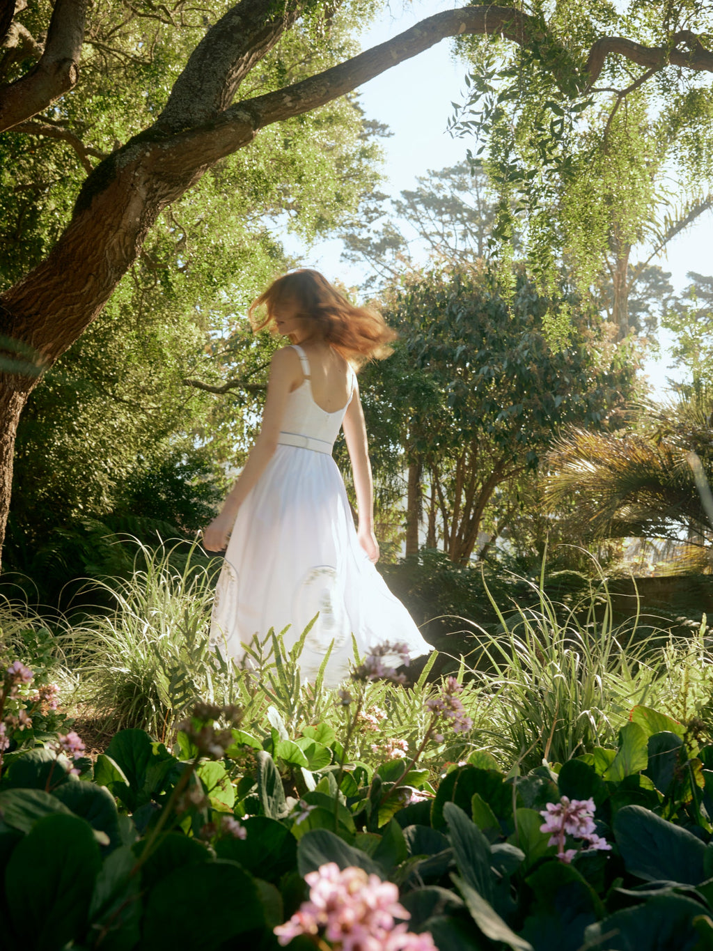 Model twirling in a white Katherine midi dress with lace embroidery and adjustable straps in a sunlit garden