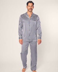A man in a navy striped pajamas set from Petite Plume stands on a plain background. The ensemble, crafted from luxurious mulberry silk, includes a long-sleeved button-up shirt and pants. His beard and short hair add to his understated elegance.