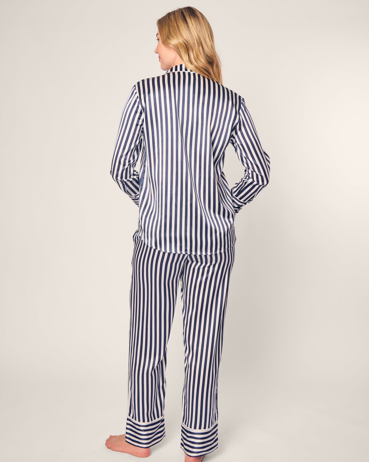 A person with long, blonde hair is wearing a Petite Plume Womens Silk Pajama Set in Navy Bengal Stripe, showcasing vertical blue and white stripes. They stand barefoot, facing away against a simple light background.