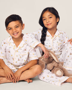 Two children in Petite Plumes Kids Twill Pajama in Easter Frolic sit on a beige floor. The boy, smiling with arms around his knees, wears white sleepwear with yellow and brown patterns. The girl with a braided hairdo holds a plush rabbit, ready for an Easter Frolic.
