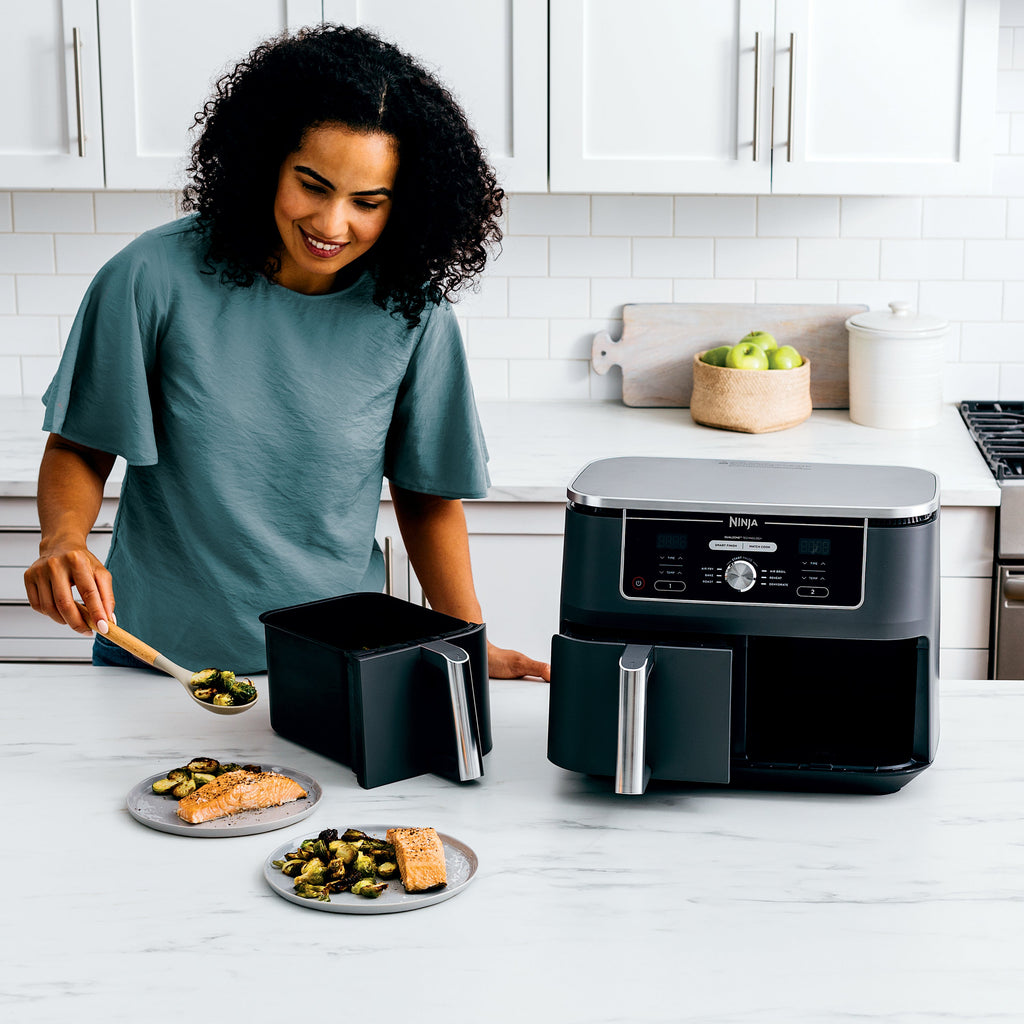 Woman serving salmon and Brussels sprouts cooked in a Ninja Foodi dual-basket air fryer on a white kitchen counter