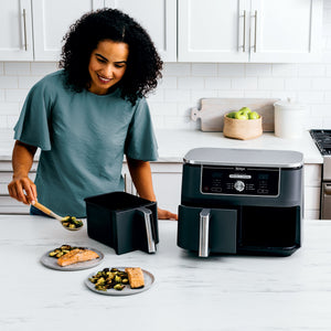 Woman serving salmon and Brussels sprouts cooked in a Ninja Foodi dual-basket air fryer on a white kitchen counter
