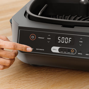 Close-up of Foodi 6-in-1 indoor grill/air fryer control panel showing digital 500F display and a finger pressing Dehydrate