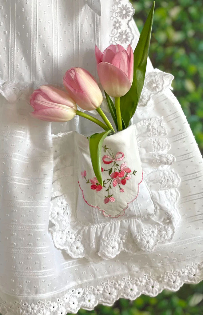 Close-up of Heirloom Handkerchief Drop-Waist Dress showcasing embroidered handkerchief pocket with pink floral applique, eyelet trim, and tulips against blurred green background