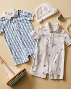 Displayed are two Petite Plume Pima cotton rompers: one with vibrant blue waves and a white collar, the other in a sailboat print. They are surrounded by a matching sailboat hat, pacifier, and wooden sailboat on a beige background.