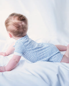 A baby with light brown hair lies on their stomach in serene comfort, wearing Petite Plumes Babys Pima Louis Play Romper in La Mer, featuring light blue wavy sea-inspired stripes, against a soft white background. The focus is on their back and head.