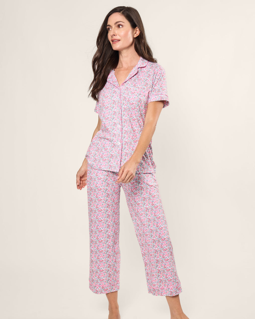 A woman wears the Womens Pima Crop Set in Fleurs de Rose by Petite Plume. Made from soft jersey knit, it features short sleeves and cropped pants. Her long dark hair frames her neutral expression against a plain backdrop, embodying the spirit of bedtime ease.