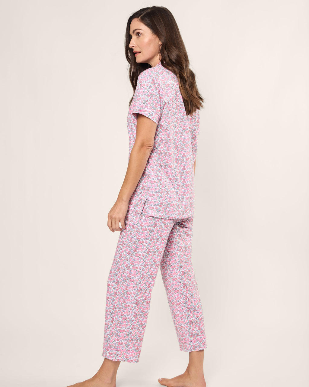 A woman with long brown hair models the Womens Pima Crop Set in Fleurs de Rose by Petite Plume, standing barefoot and gazing to the side against a plain light backdrop, capturing a serene bedtime vibe.