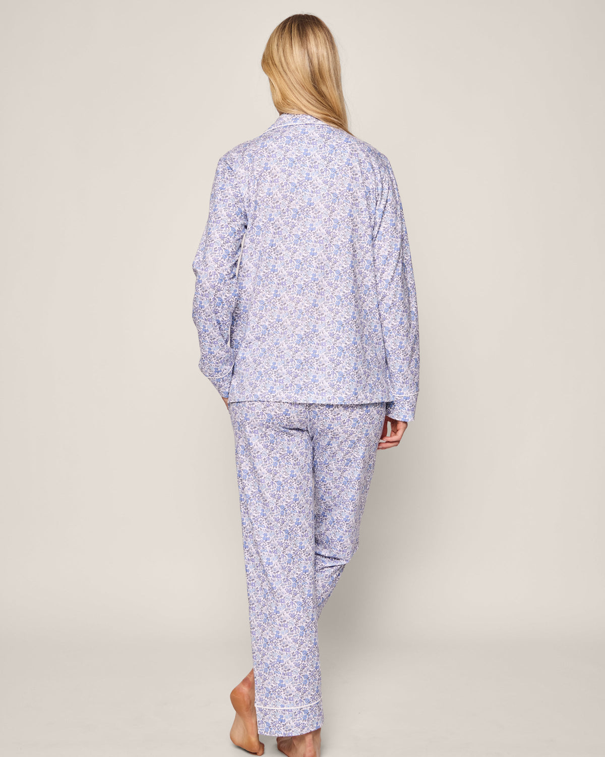 A person with long blonde hair stands barefoot, facing away, wearing the vintage-inspired Petite Plume Womens Pima Pajama in Fleur dAzur. The light blue patterned pajamas with long sleeves and pants create an elegant look against a plain background.