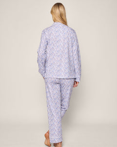 A person with long blonde hair stands barefoot, facing away, wearing the vintage-inspired Petite Plume Womens Pima Pajama in Fleur dAzur. The light blue patterned pajamas with long sleeves and pants create an elegant look against a plain background.