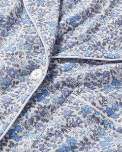 Close-up of Petite Plumes Womens Pima Pajama in Fleur dAzur, featuring a floral pattern in blue and purple on luxurious Peruvian Pima cotton. A white button, piping, and visible pocket enhance its vintage-inspired look against a soft grayish background.