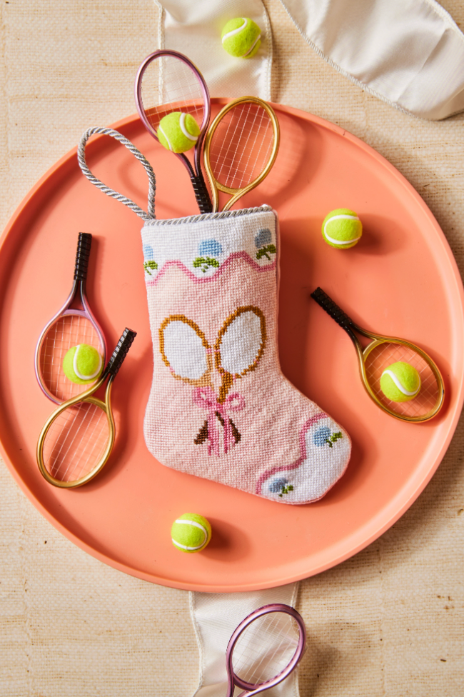 Top view of pink needlepoint stocking ornament with tennis-racket motif and braided loop on coral plate with mini rackets and tennis balls