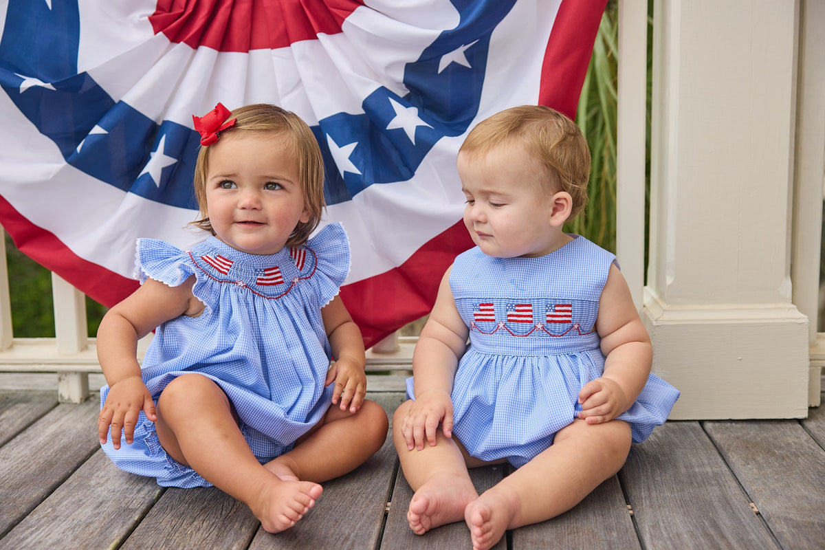 Little English traditional children's clothing.  Flutter sleeve bubble in royal blue mini check fabric and smocking with American flag bunting for baby girls