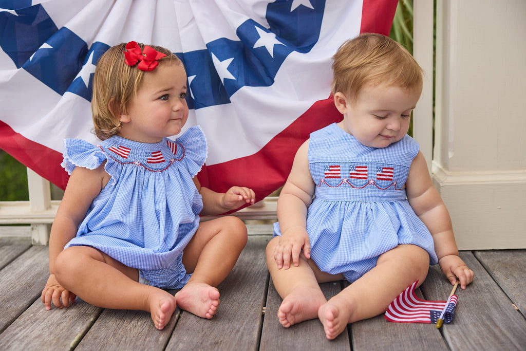 Little English traditional children's clothing.  Flutter sleeve bubble in royal blue mini check fabric and smocking with American flag bunting for baby girls