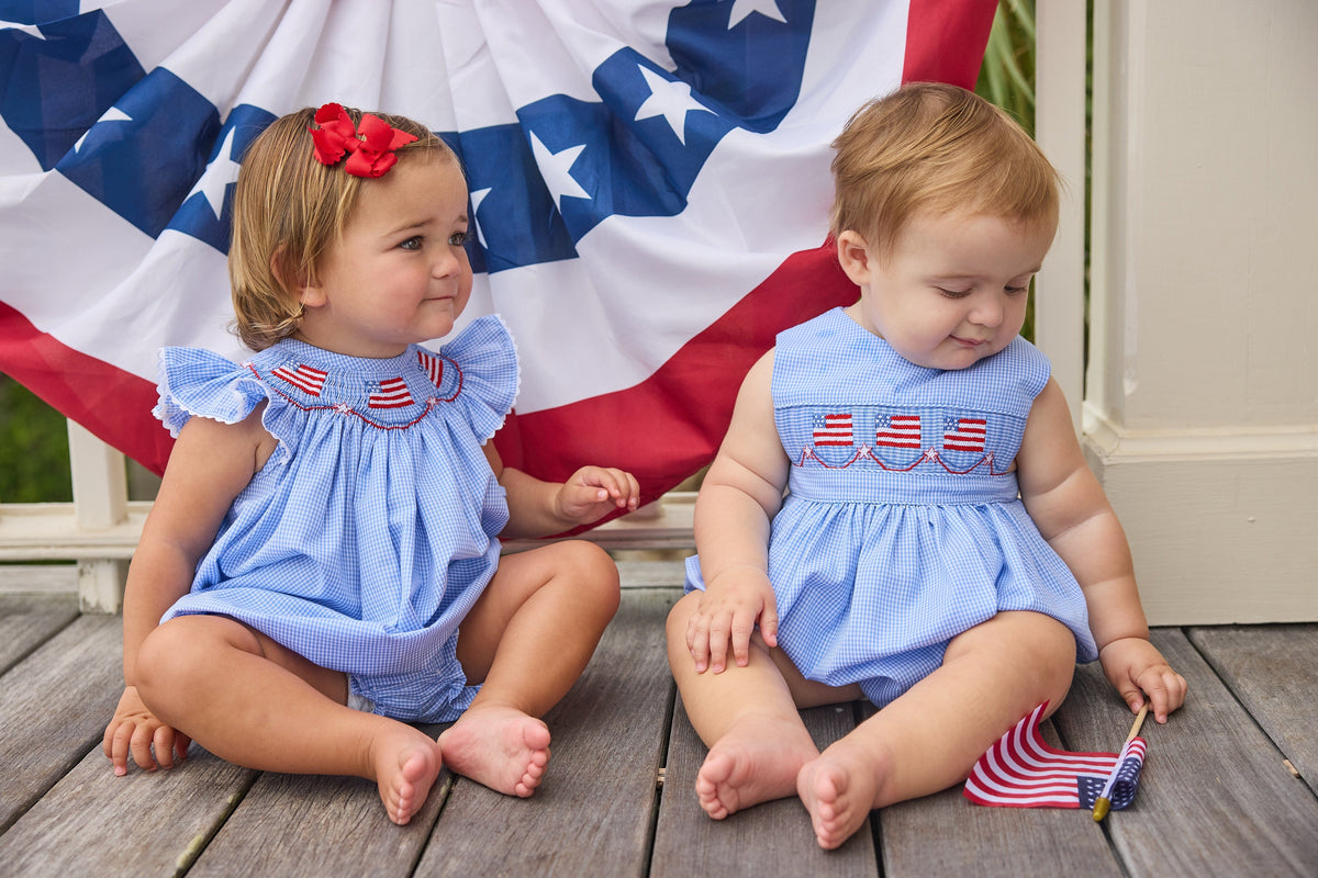 Little English traditional children's clothing.  Flutter sleeve bubble in royal blue mini check fabric and smocking with American flag bunting for baby girls