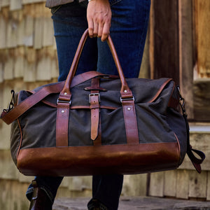 Person holding a Canvas Weekender Duffel Bag by Tom Beckbe, featuring brown leather accents, in front of a wooden building.