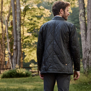 Man in a Tom Beckbe Paddock Quilted Jacket with breathable insulation stands in forest, sunlight filtering through leaves.