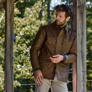 A man in a Tom Beckbe Paddock Quilted Jacket leans against a post outdoors, with trees in the background.