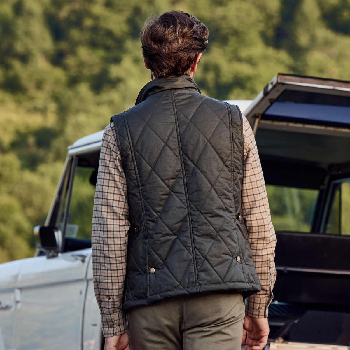 A person in a Tom Beckbe Paddock Quilted Vest stands by a vehicle amid trees, kept warm by HD Wool.