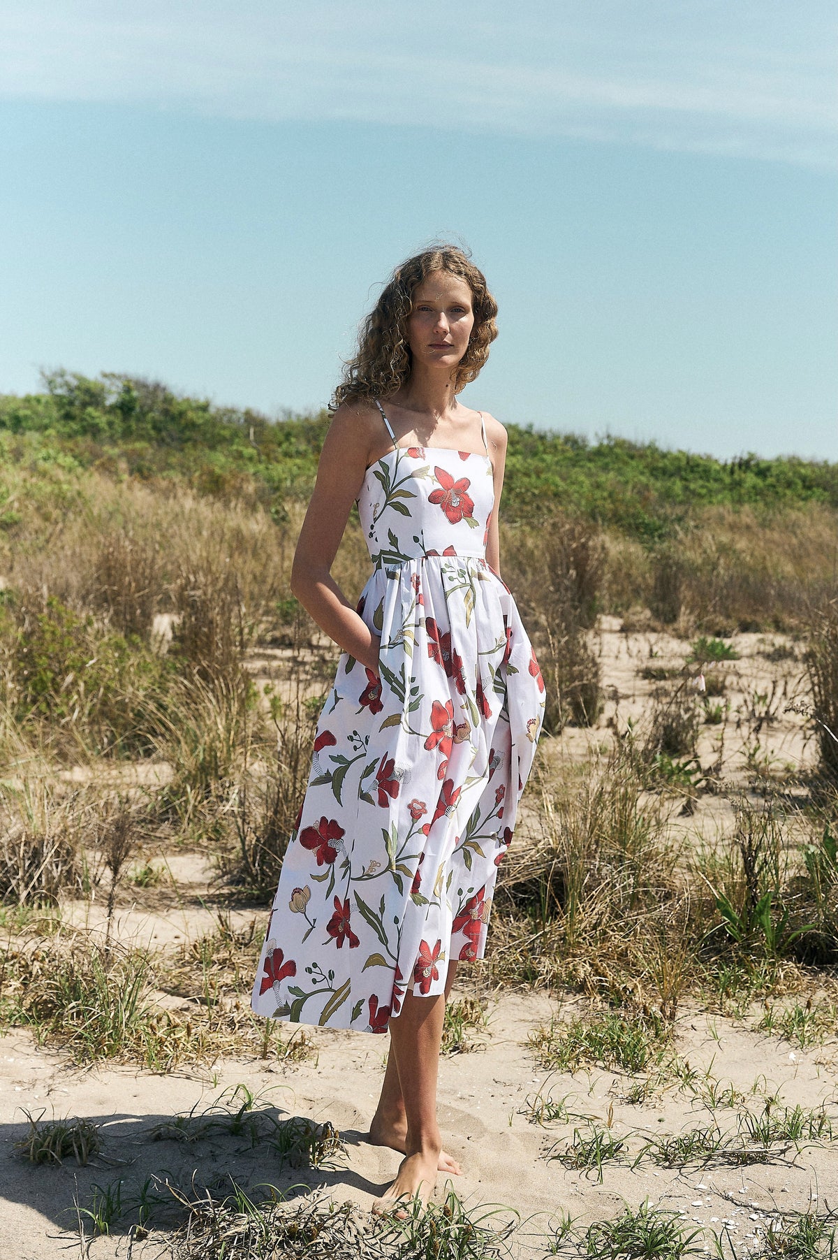 Woman wearing red hibiscus floral skinny-strap poof dress with fitted bodice and pockets standing barefoot on a sandy beach