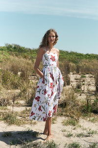 Woman wearing red hibiscus floral skinny-strap poof dress with fitted bodice and pockets standing barefoot on a sandy beach