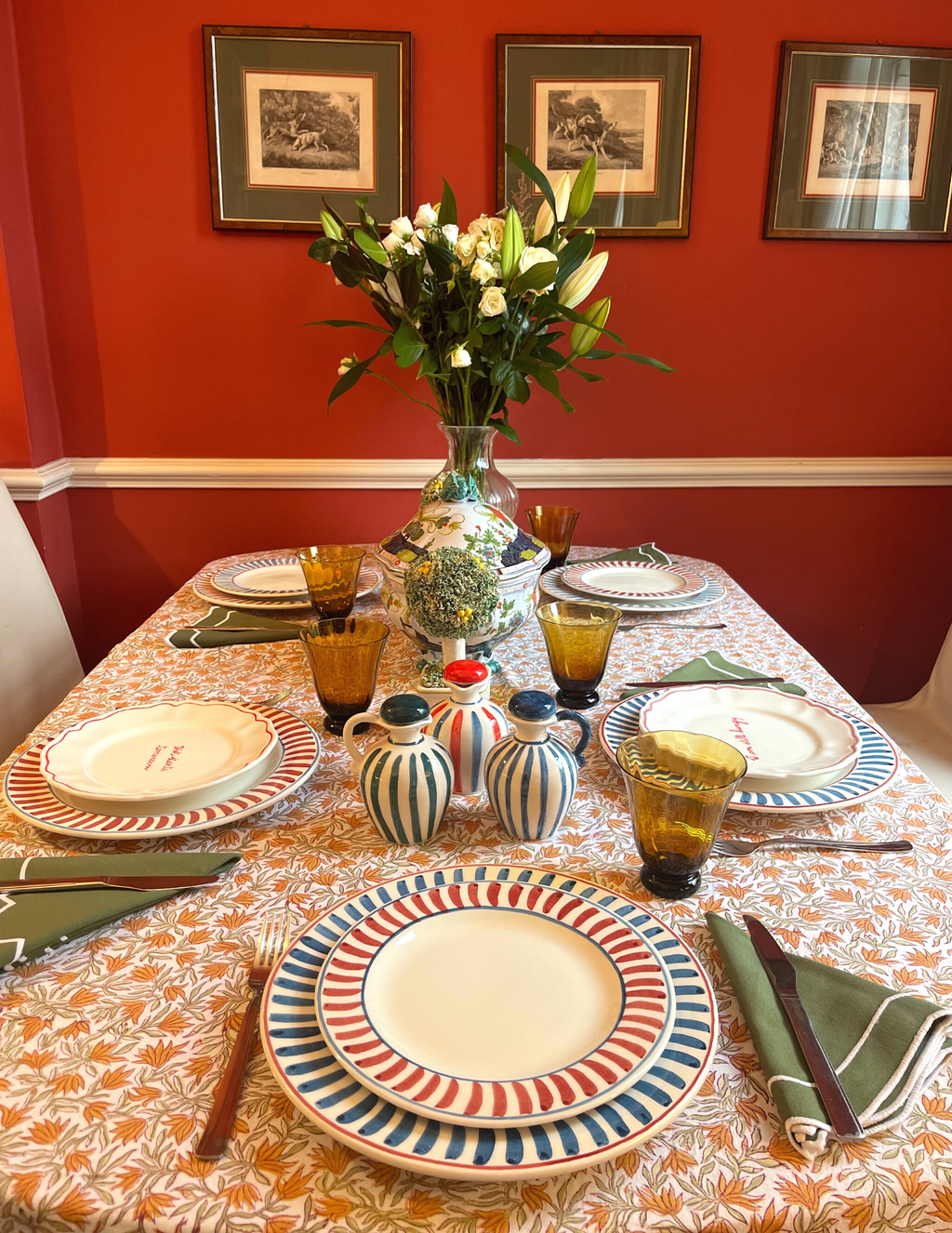 Dining table set with scalloped plates with red and blue rims, amber glasses, green napkins, and flower vase against red wall