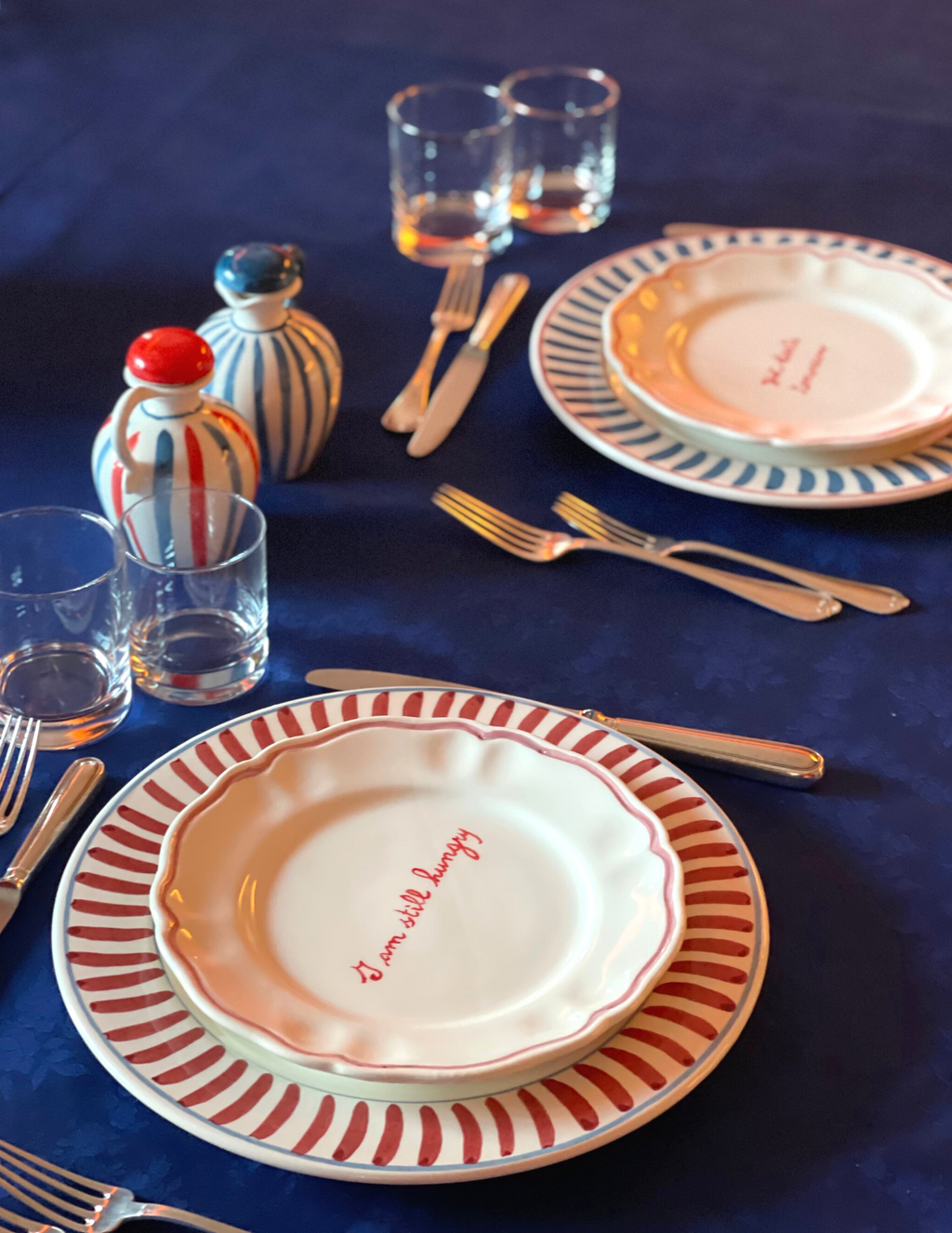 Angled view of scalloped ceramic plate with red handwritten I am still hungry text on red-striped charger over dark tablecloth