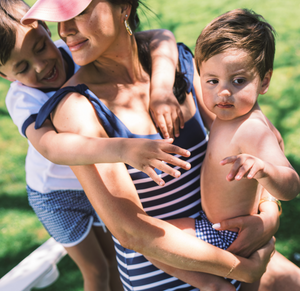 Woman wearing navy Breton-striped tie-knot one-piece swimsuit holding two young children outdoors on grass