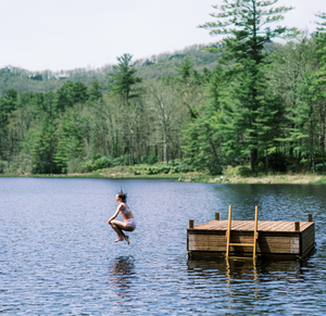 Woman jumping off a wooden dock into a lake wearing raspberry red high-waist bikini bottom, forested shoreline in background