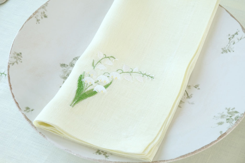 Top-down view of a folded light-green linen napkin with embroidered lily-of-the-valley sprig on a floral dinner plate