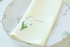 Top-down view of a folded light-green linen napkin with embroidered lily-of-the-valley sprig on a floral dinner plate