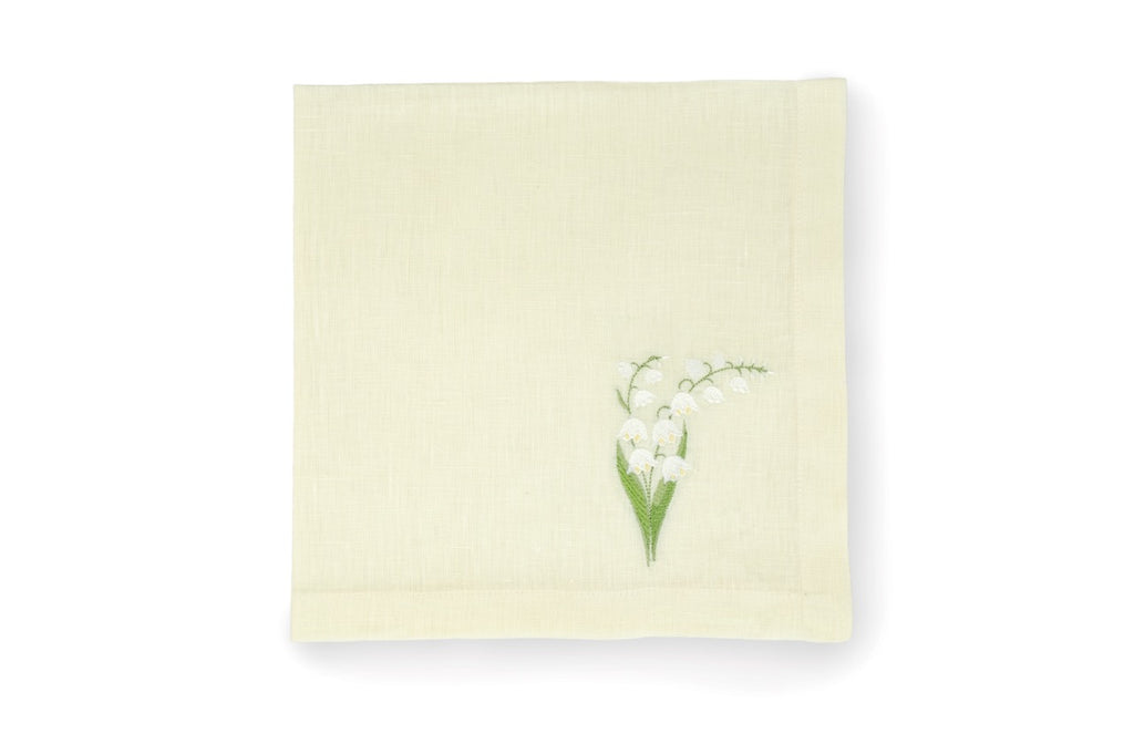 Top down view of a pale green linen napkin folded showing an embroidered lily of the valley sprig at the corner on a white background